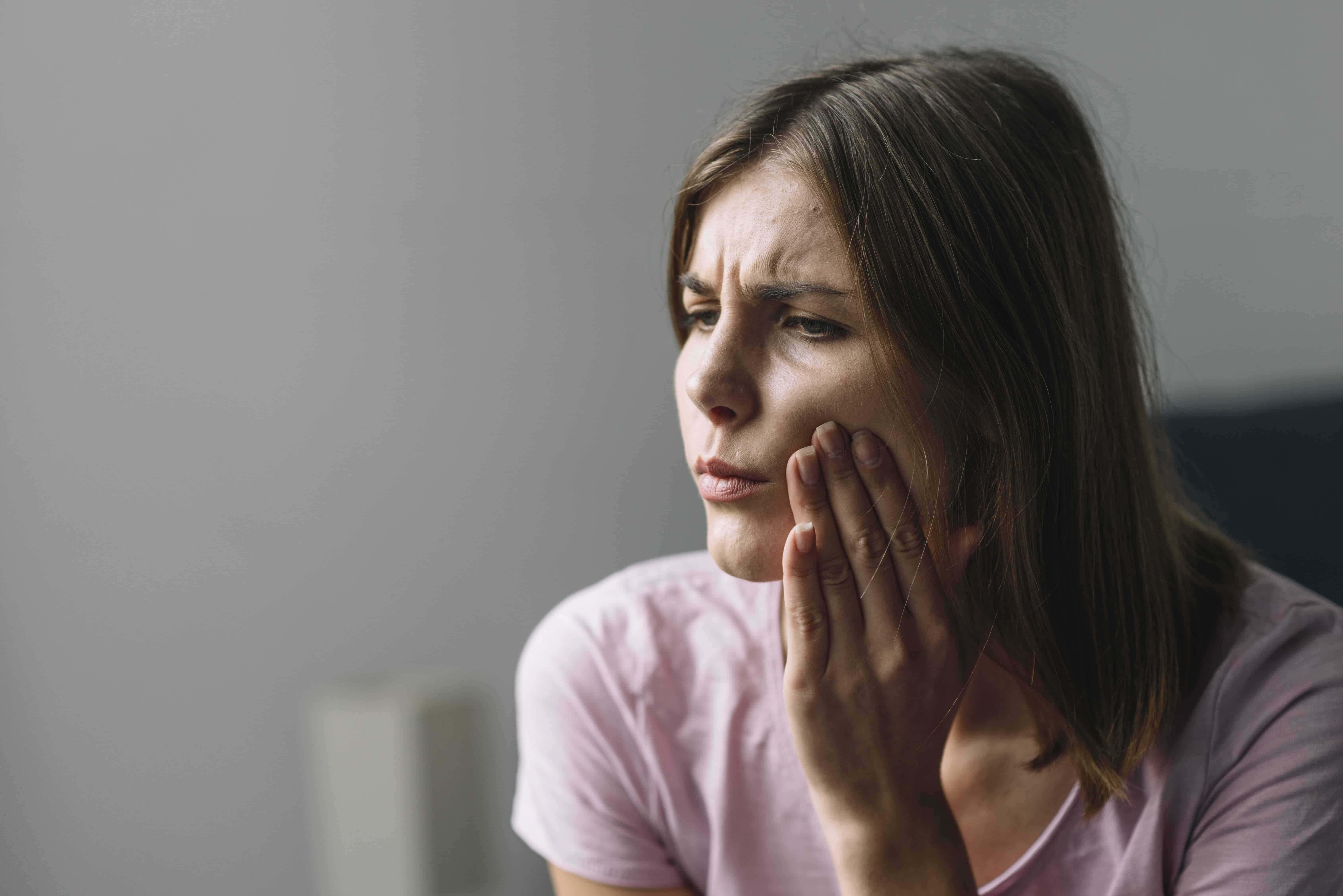 Emergency Dentist Raynes Park - A Young Woman Is Shown In Visible Discomfort, Holding Her Jaw, Indicative Of Severe Tooth Pain. This Image Emphasizes The Urgent Need For Dental Care, Such As That Provided By Beverley Dental In Raynes Park. The Setting Suggests A Home Environment, Highlighting The Importance Of Accessible Emergency Dental Services. The Expression On Her Face Underlines The Urgency And Distress That Dental Emergencies Can Cause, Showcasing The Essential Services Offered By Beverley Dental To Alleviate Pain And Provide Immediate Relief.